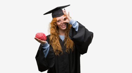 Young redhead student woman wearing graduated uniform holding brain with happy face smiling doing ok sign with hand on eye looking through fingers