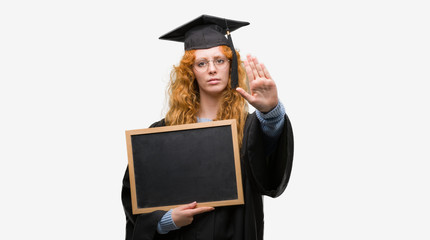 Young redhead student woman wearing graduated uniform holding blackboard with open hand doing stop sign with serious and confident expression, defense gesture