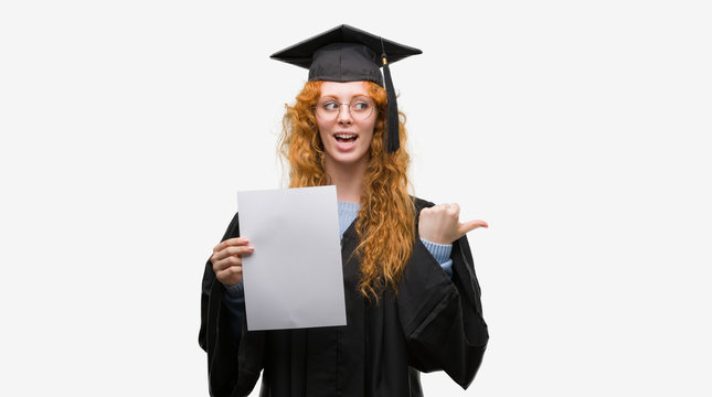 Young redhead woman wearing graduate uniform holding degree pointing and showing with thumb up to the side with happy face smiling