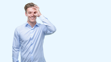 Young handsome blond man with happy face smiling doing ok sign with hand on eye looking through fingers