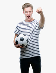 Young handsome blond man holding soccer ball annoyed and frustrated shouting with anger, crazy and yelling with raised hand, anger concept