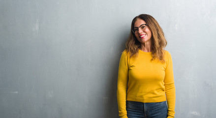 Middle age hispanic woman over grey wall wearing glasses looking away to side with smile on face,...
