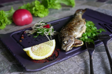 Grilled fish on a dark platter with herbs and lemon. Served with vegetables.