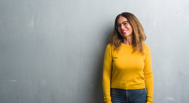 Middle age hispanic woman over grey wall wearing glasses looking away to side with smile on face, natural expression. Laughing confident.