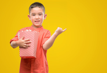 Dark haired little child eating popcorn very happy pointing with hand and finger to the side