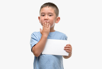 Dark haired little child holding a blank card cover mouth with hand shocked with shame for mistake, expression of fear, scared in silence, secret concept