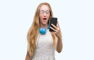 Fototapeta premium Blonde teenager woman holding smartphone and wearing headphones scared in shock with a surprise face, afraid and excited with fear expression