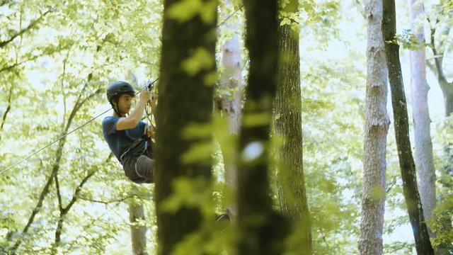 Person slide down the zip-line in slow motion 4K. Long shot rack focus tracking of a climber in focus sliding on a wire while wearing safety equipment and helmet. Climbing experience in adventure park