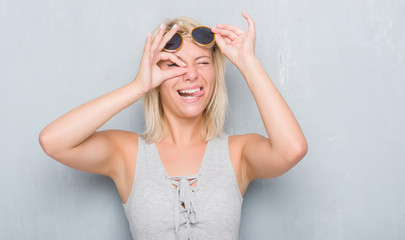 Adult caucasian woman over grunge grey wall wearing retro sunglasses with happy face smiling doing ok sign with hand on eye looking through fingers