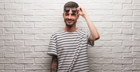 Young adult man wearing sunglasses standing over white brick wall winking looking at the camera...