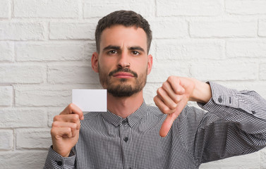 Young adult man over brick wall holding blank card with angry face, negative sign showing dislike...