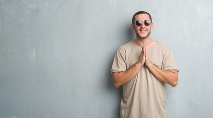 Young caucasian man over grey grunge wall wearing sunglasses praying with hands together asking for forgiveness smiling confident.