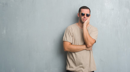 Young caucasian man over grey grunge wall wearing sunglasses thinking looking tired and bored with depression problems with crossed arms.