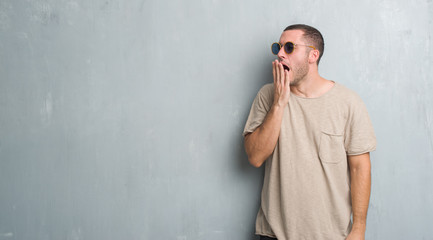 Young caucasian man over grey grunge wall wearing sunglasses bored yawning tired covering mouth with hand. Restless and sleepiness.