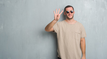 Young caucasian man over grey grunge wall wearing sunglasses showing and pointing up with fingers number four while smiling confident and happy.