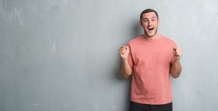 Young caucasian man over grey grunge wall celebrating surprised and amazed for success with arms raised and open eyes. Winner concept.
