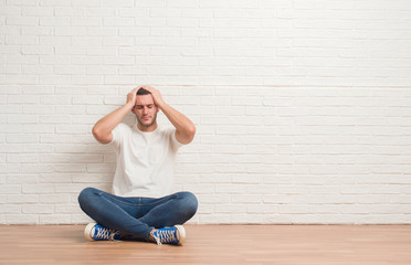Young caucasian man sitting on the floor over white brick wall suffering from headache desperate and stressed because pain and migraine. Hands on head.