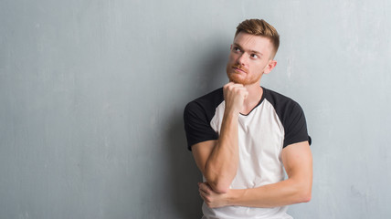 Young redhead man over grey grunge wall with hand on chin thinking about question, pensive expression. Smiling with thoughtful face. Doubt concept.