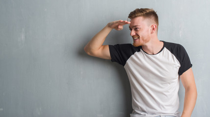 Young redhead man over grey grunge wall very happy and smiling looking far away with hand over head. Searching concept.