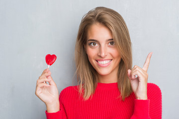 Beautiful young woman over grunge grey wall eating red heart lollipop candy surprised with an idea or question pointing finger with happy face, number one