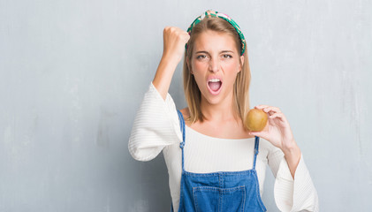 Beautiful young woman over grunge grey wall holding fresh kiwi annoyed and frustrated shouting with anger, crazy and yelling with raised hand, anger concept