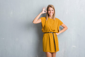 Fototapeta premium Beautiful young woman standing over grunge grey wall wearing a dress smiling positive doing ok sign with hand and fingers. Successful expression.
