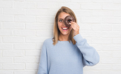 Beautiful young woman over white brick wall eating chocolate donut with a happy face standing and smiling with a confident smile showing teeth