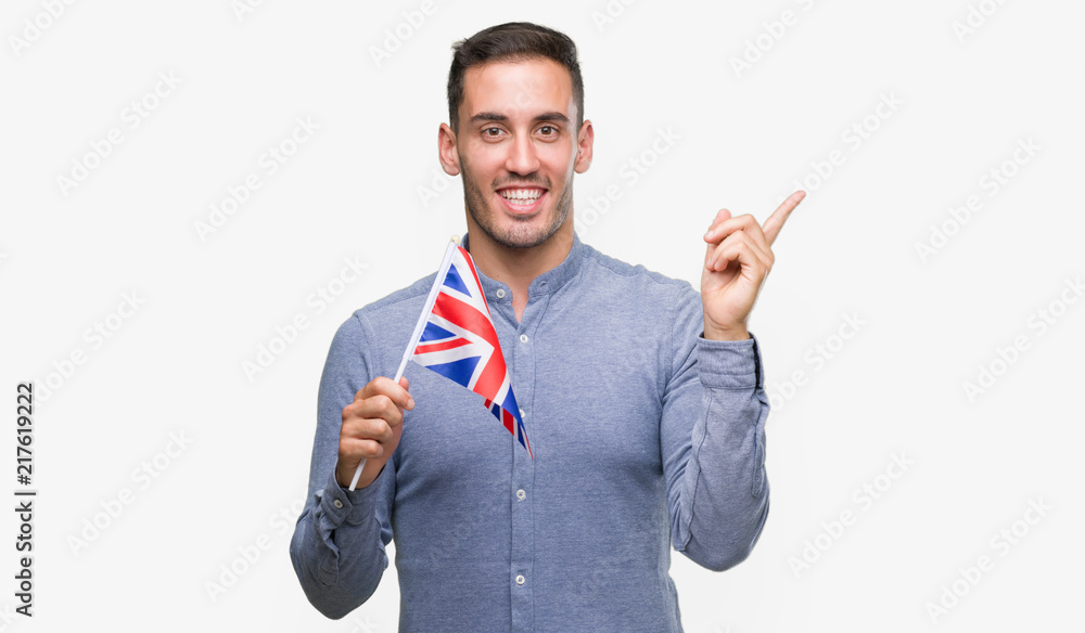 Poster handsome young man holding a flag of united kingdom very happy pointing with hand and finger to the 