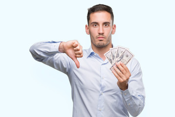 Handsome young man holding money with angry face, negative sign showing dislike with thumbs down,...