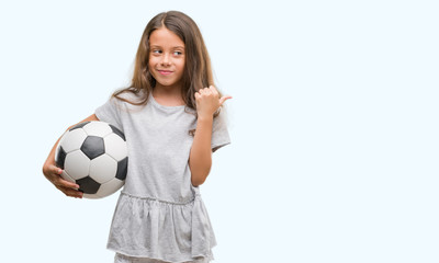 Brunette hispanic girl holding soccer football ball pointing and showing with thumb up to the side with happy face smiling