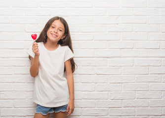 Young hispanic kid over white brick wall eating red heart lollipop candy with a happy face standing and smiling with a confident smile showing teeth