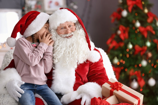 Little girl whispering in authentic Santa Claus' ear indoors