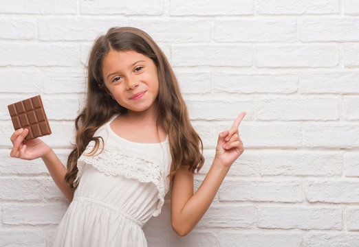 Young Hispanic Kid Over White Brick Wall Eating Chocolate Bar Very Happy Pointing With Hand And Finger To The Side