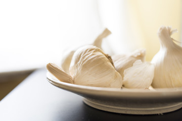 Heads of hard neck Californian garlic on white ceramic bowl.  Bright window behind black table top.