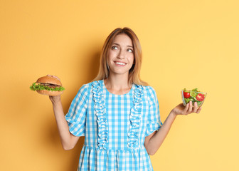 Young woman holding burger and salad on color background. Choice between diet and unhealthy food