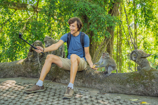 Selfie with monkeys. Young man uses a selfie stick to take a photo or video blog with cute funny monkey. Travel selfie with wildlife in Bali
