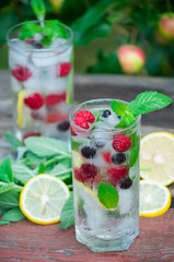 Cold lemonade with lemon, mint and berries in a transparent glass with facets. On an old wooden table. In the background, the green foliage is a soft blurred focus.