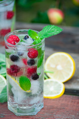 Cold lemonade with lemon, mint and berries in a transparent glass with facets. On an old wooden table. In the background, the green foliage is a soft blurred focus.