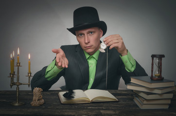 Man in bowler hat and suit is sitting by table and showing a pocket watch in his hand. Late for a...