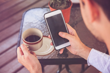 Attractive man using cellphone and drinking coffee while sitting on the tropical cafe. Focus on phone.
