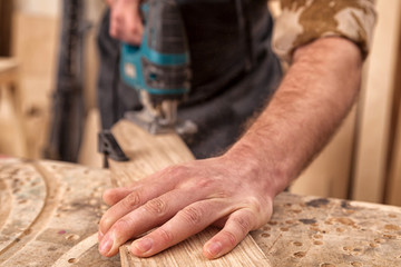 Close up of experienced carpenter in work clothes and small buiness owner  carpenter saw and processes the edges of a wooden bar with a jig saw  in a light workshop