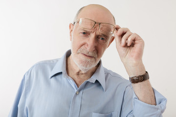 Portrait of attractive European mature bearded male in his sixties raising rectangular spectacles and staring at camera with scrutinized skeptical look, his eyes expressing disbelief and suspicion