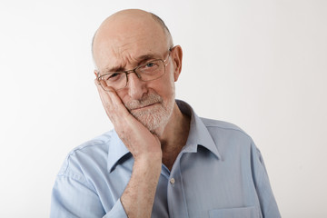 Portrait of frustrated upset mature male in his sixties holding hand on his cheek while suffering from terrible toothache. Retired elderly Caucasian man with gray beard having pain in teeth