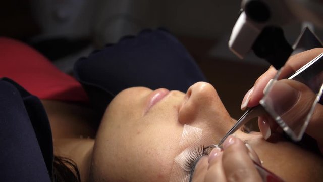 Woman Eye with Long Eyelashes. Eyelash Extension. Lashes, close up, selected focus. Gluing artificial eyelashes with tweezers. Beautician in magnifier glasses performs laborious small work.