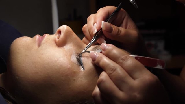Woman eye with long eyelashes. Eyelash extension. Gluing artificial eyelashes with tweezers. A woman lies under a lamp on a cosmetic procedure. movement of the camera.