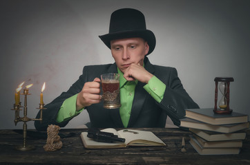 Severe detective agent man in a bowler hat is sitting by the table and drinking a tea from cup....