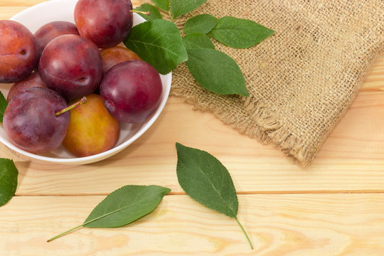 Plums in white bowl and leaves on wooden surface closeup