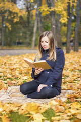 Young pregnant girl reads a book sitting on a plaid in an autumn