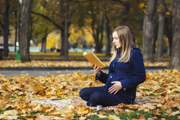 Young pregnant girl reads a book sitting on a plaid in an autumn