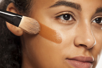 portrait of a young dark-skinned woman applying liquid makeup base with brush on her face on a...